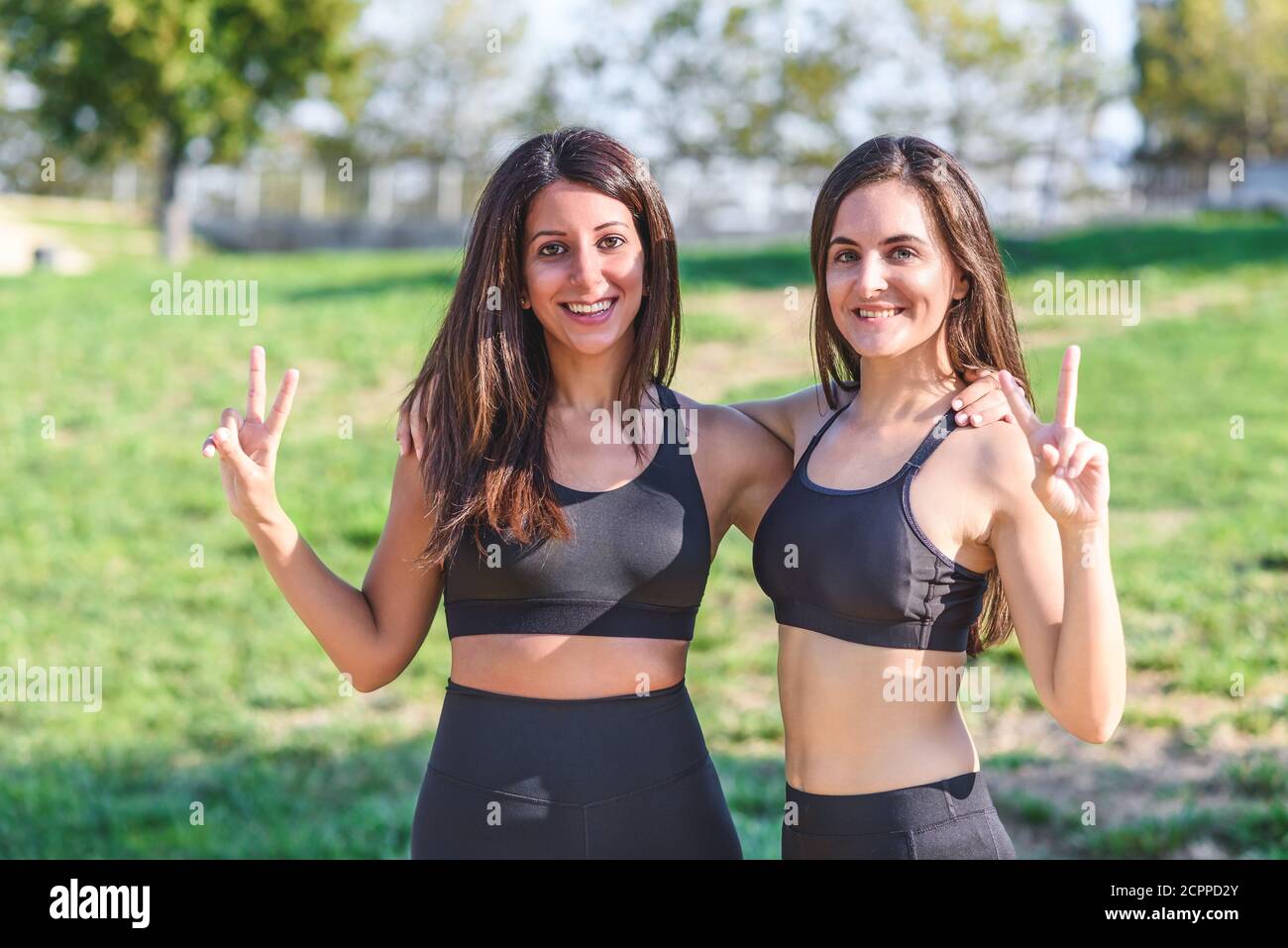 kaukasische und lateinische Freundinnen kleiden schwarze Sportkleidung umarmt Und die Geste des Sieges mit den Fingern zu tun Stockfoto