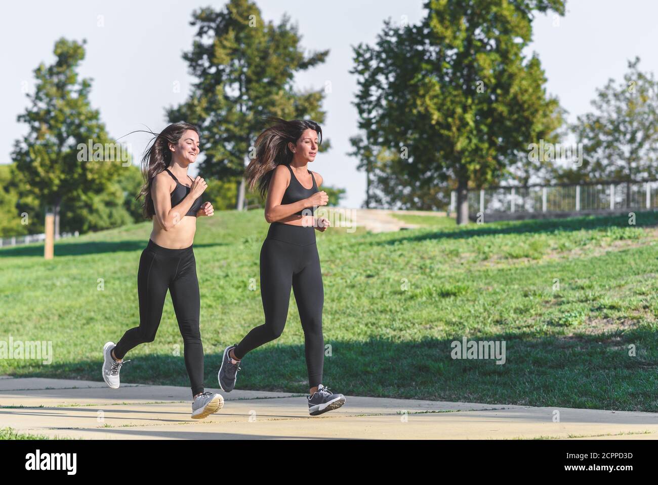 kaukasische und lateinische Freundinnen mit schwarzer Sportbekleidung laufen Gemeinsam in einem grünen Park Stockfoto