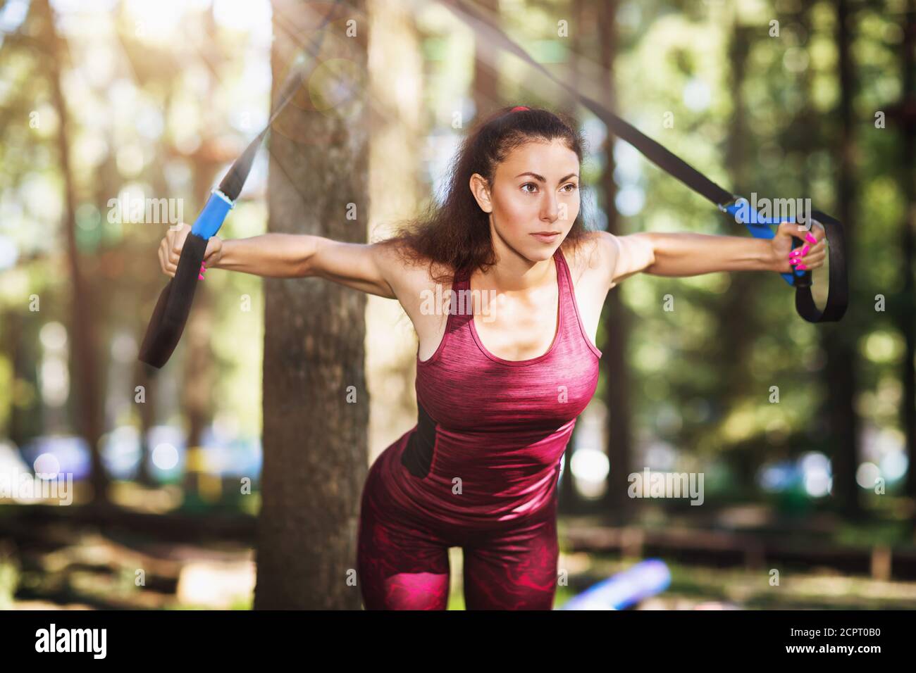 Junge Frau im Trainingsanzug macht Übungen im Park mit Fitness-Riemen an einem Baum befestigt. Der Athlet trainiert die Brustmuskeln Stockfoto