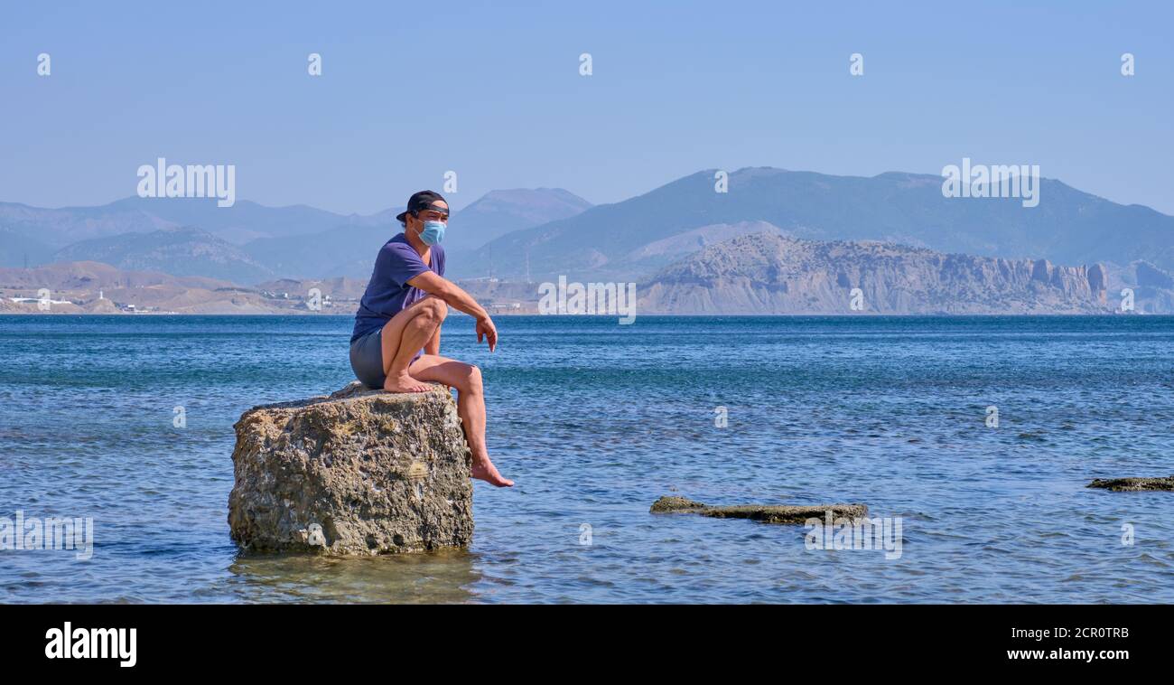 Senior Asian Mann in Gesichtsmaske, Shorts, T-Shirt und Baseballmütze sitzt allein auf einem Stein in einer Meeresbucht vor einem Hintergrund von Bergen Banner Stockfoto