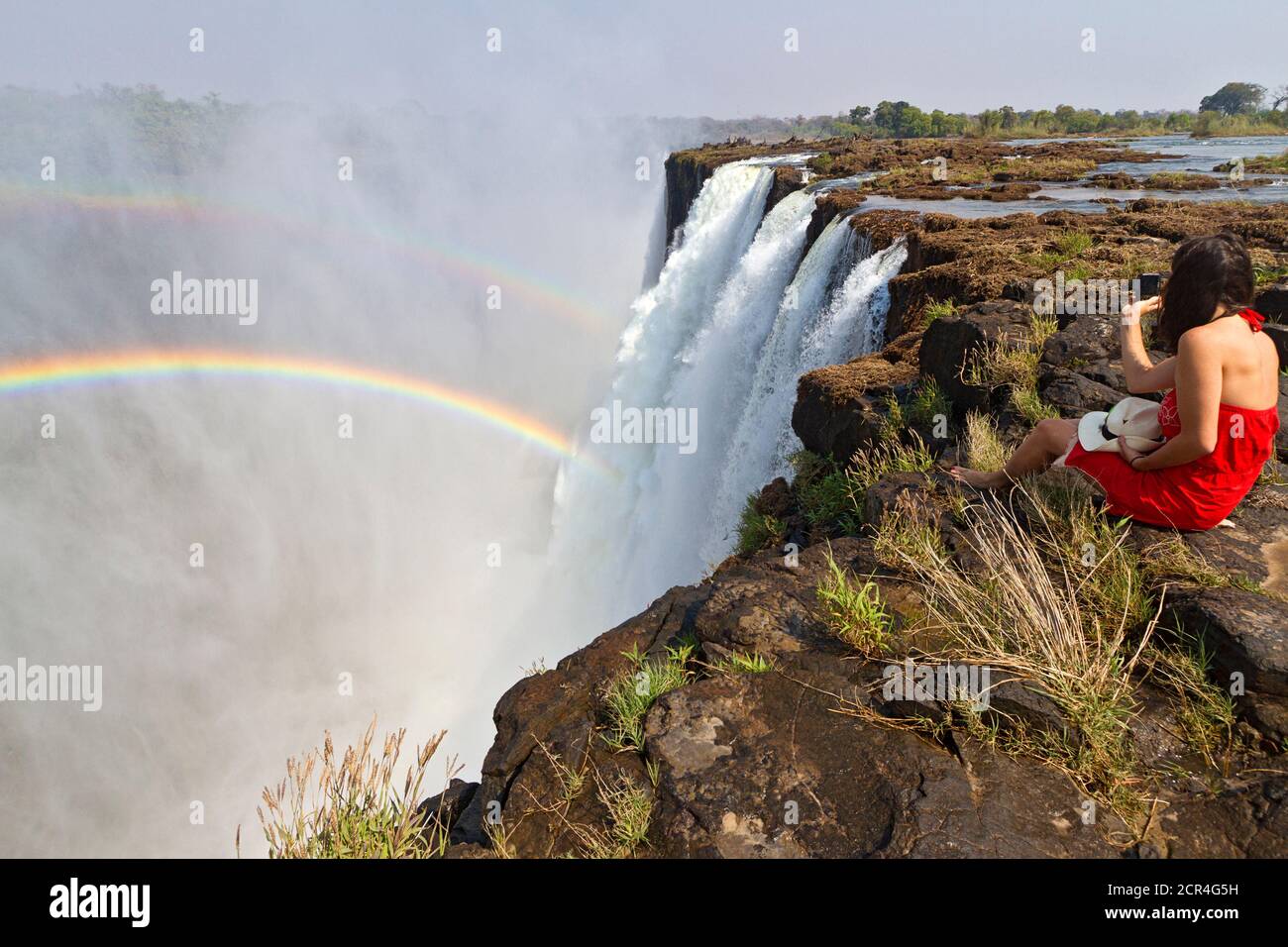 Die junge Frau sitzt am Rande der Victoria Falls, um den doppelten Regenbogen in Livingstone, Sambia, Afrika, zu sehen und zu fotografieren. Victoria Falls ist fast eine Meile breit und 360 Fuß tief und aus der Luft, sieht aus wie die Erde in zwei gerissen wurde. Sambia, Afrika. Stockfoto