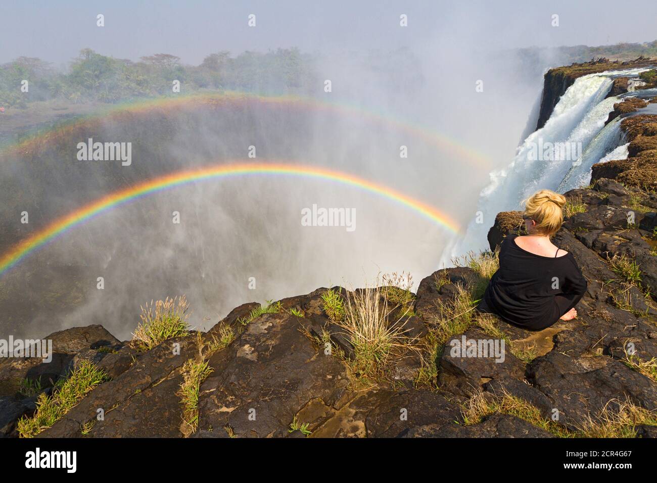 Die junge Frau sitzt am Rande der Victoria Falls, um den doppelten Regenbogen in Livingstone, Sambia, Afrika, zu sehen und zu fotografieren. Victoria Falls ist fast eine Meile breit und 360 Fuß tief und aus der Luft, sieht aus wie die Erde in zwei gerissen wurde. Sambia, Afrika. Stockfoto