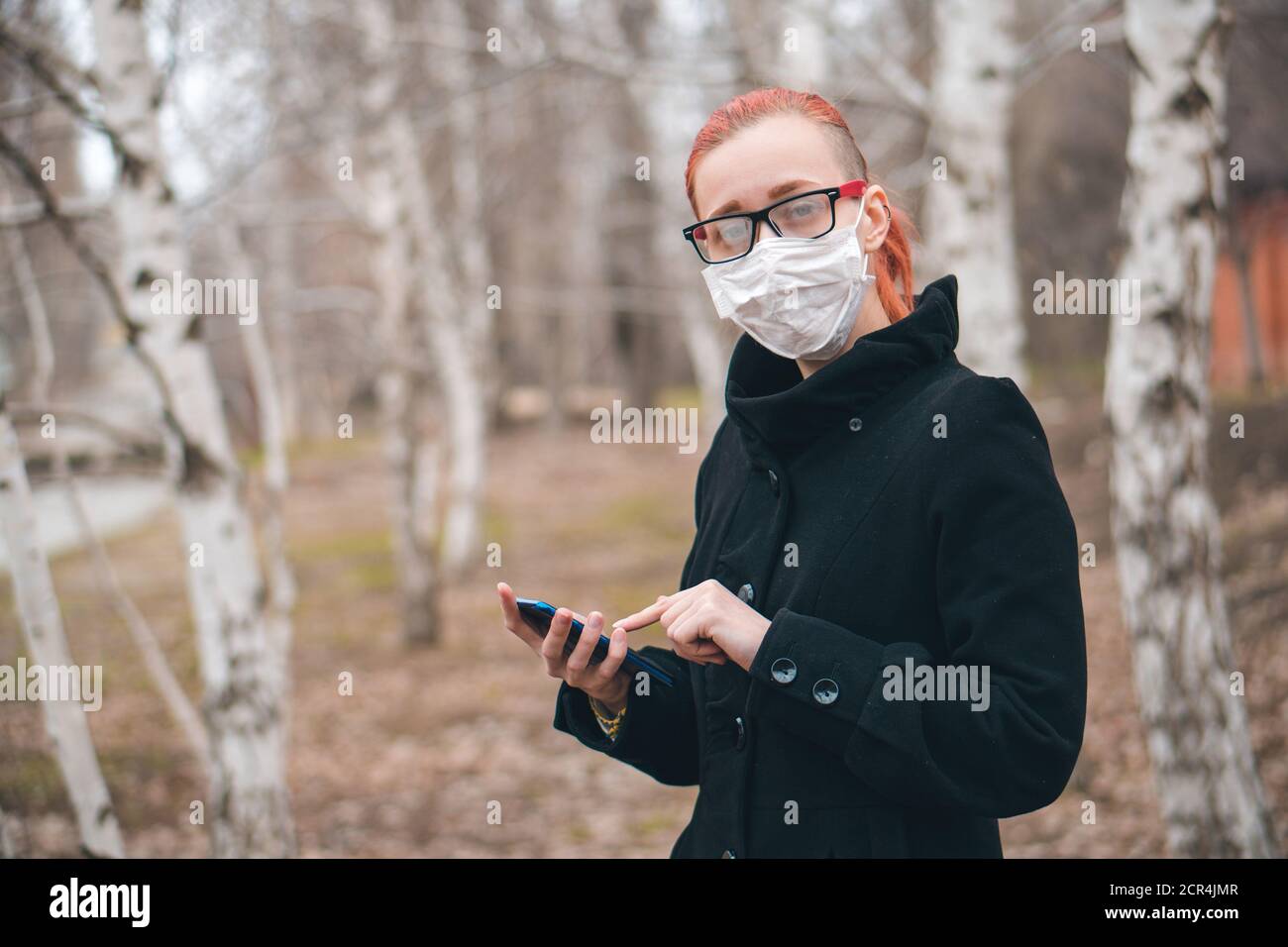 Junge schöne Frau in medizinische Maske hält Telefon in der Hand im Park und schaut auf Kamera. Health Care oder Epidemie Konzept mit Kopieplatz. Stockfoto