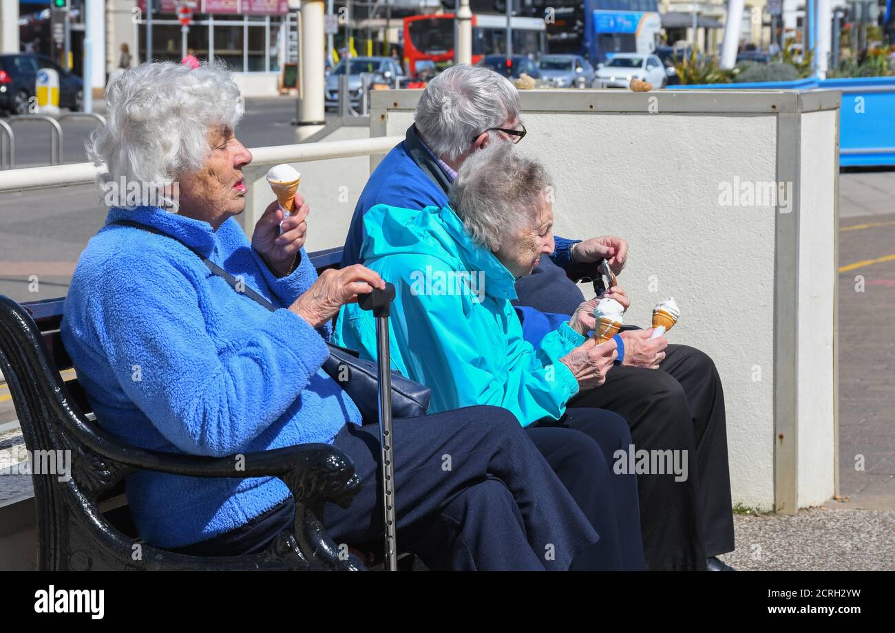 Eine kleine Gruppe älterer Menschen, die draußen auf einer Bank sitzen und Eis essen, im Frühjahr in England. Alte ältere Leute essen draußen Eis. Stockfoto