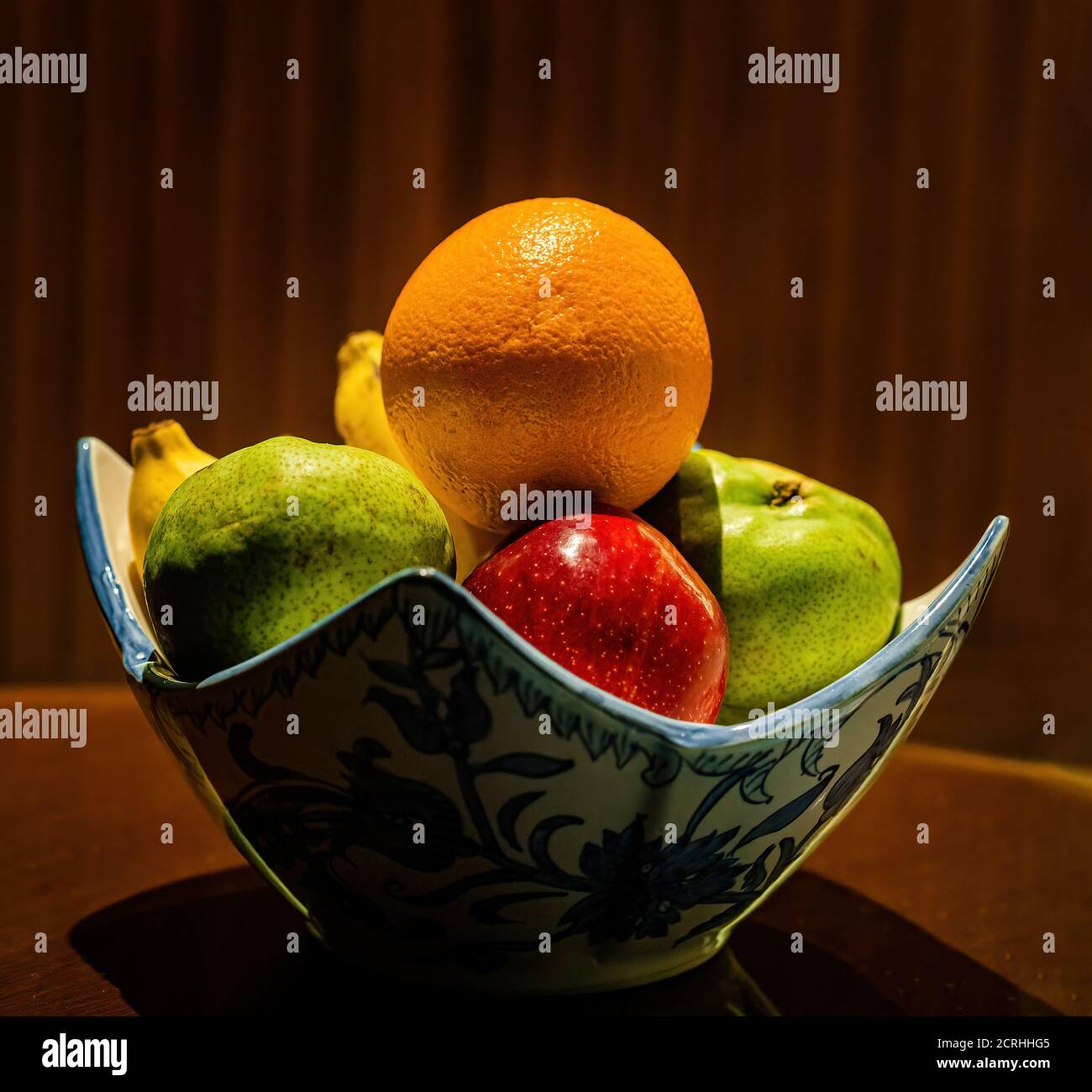 Eine Schüssel mit frischem Obst, darunter Apfel, Orange, Birne und Banane, auf einem Tisch mit Beleuchtung von oben Stockfoto