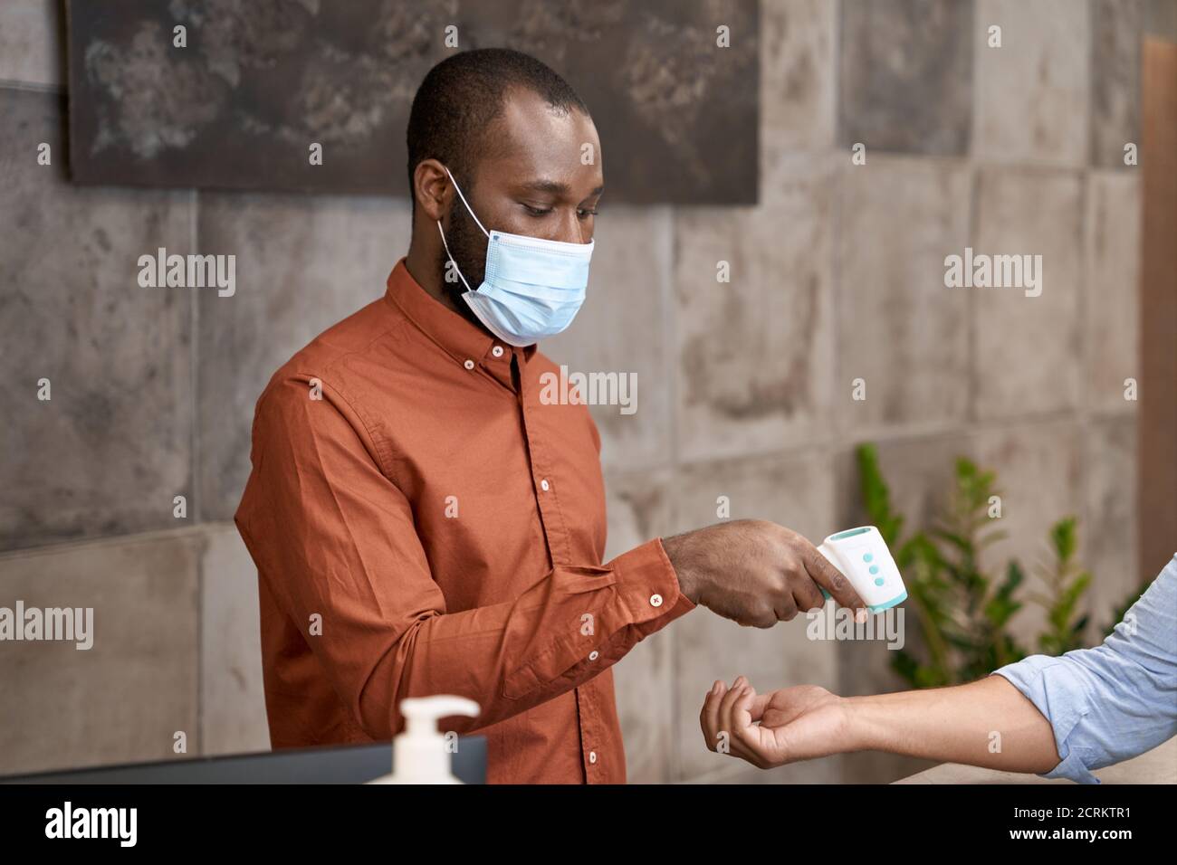 Afrikanischer männlicher Rezeptionist trägt medizinische Maske mit Infrarot-Thermometer für Körpertemperatur des Büroangestellten messen Stockfoto