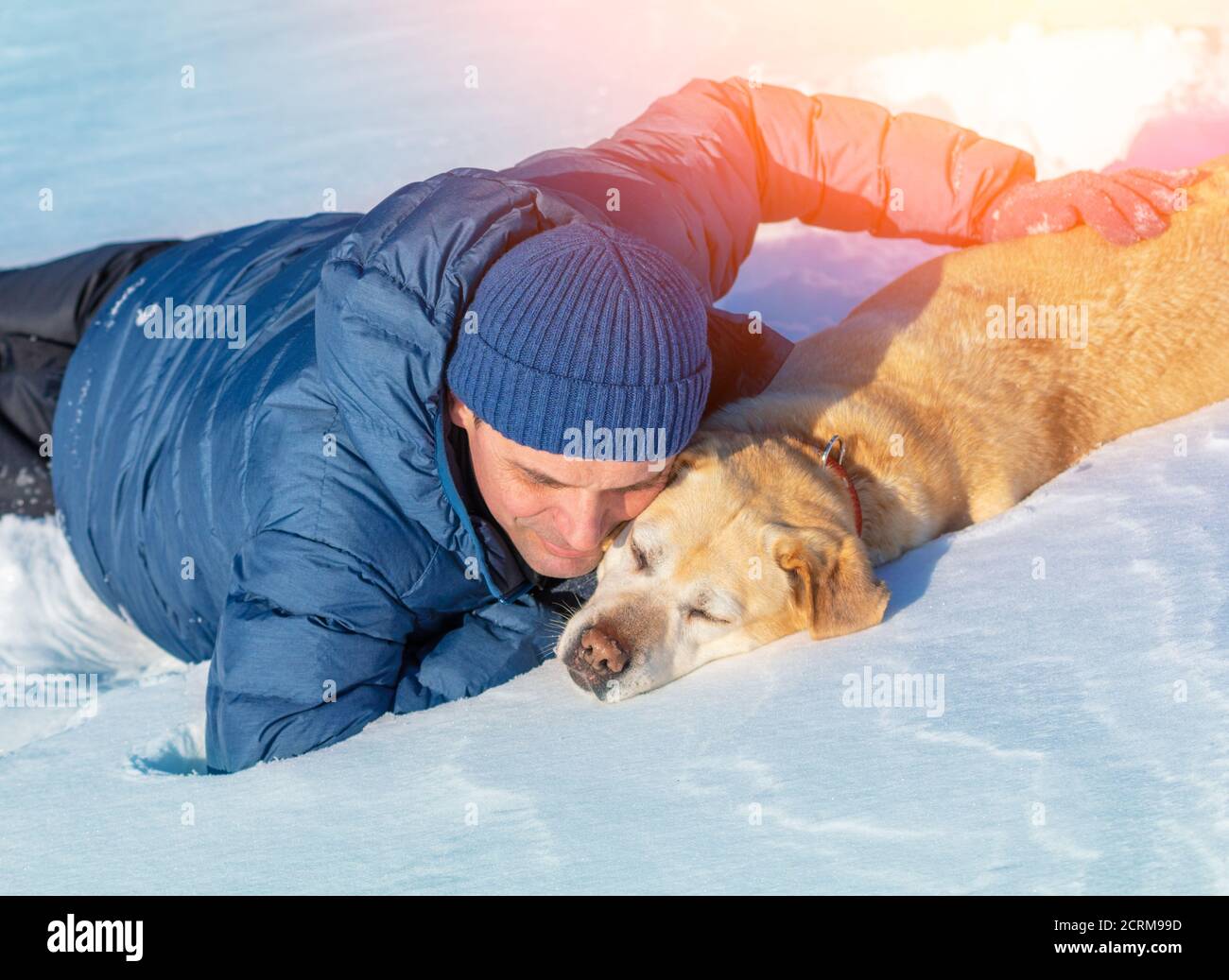 Ein glücklicher lächelnder Mann mit einem Labrador Retriever Hund liegt Im Schnee im Winter Stockfoto