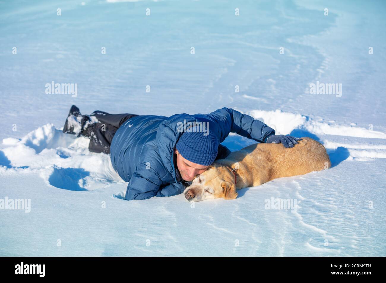 Ein glücklicher lächelnder Mann mit einem Labrador Retriever Hund liegt Im Schnee im Winter Stockfoto
