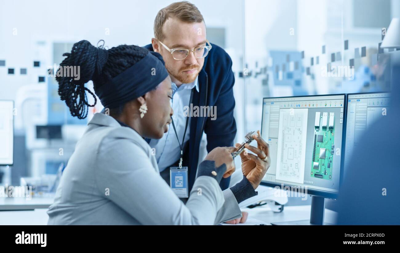Moderne Elektronikfabrik: Weibliche Vorgesetzte spricht mit einem männlichen Elektroingenieur, der auf Computer mit CAD-Software arbeitet. Design PCB, Mikrochips Stockfoto