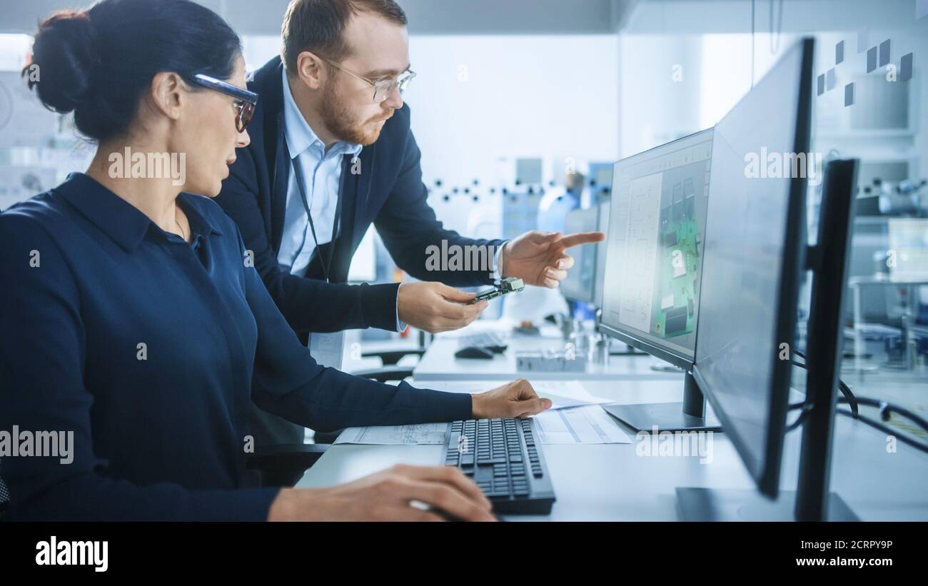 Moderne Elektronikfabrik: Männlicher Vorgesetzter spricht mit einer weiblichen Elektroingenieurin, die auf Computer mit CAD-Software arbeitet. Entwicklung von Leiterplatten, Mikrochips Stockfoto