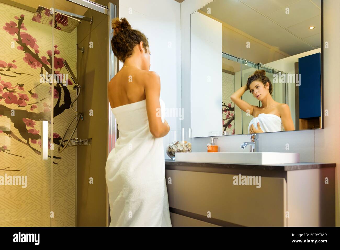 Junge Frau eingewickelt in ein sauberes weißes Handtuch stehend suchen Bei sich im Badezimmerspiegel mit der Hand zu Ihr Log-Haar, als sie sich auf eine Show vorbereitet Stockfoto