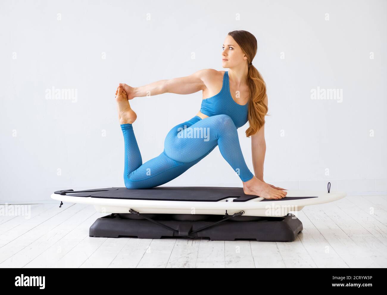 Junge Frau macht Dehnübungen der Oberschenkelmuskulatur ihres hinteren Beins Muskeln auf einem Surfset in einer Seitenansicht in einem High-Key-Fitness-Studio in einem Gesundheits-und Fitness-Co Stockfoto
