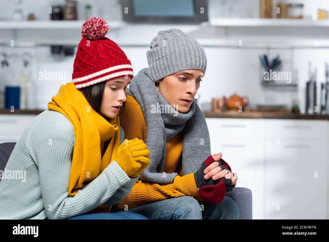 Junges Paar in warmen Hüten, Schals und Handschuhen sitzend mit geballten Händen, während sie in der kalten Küche friert Stockfoto