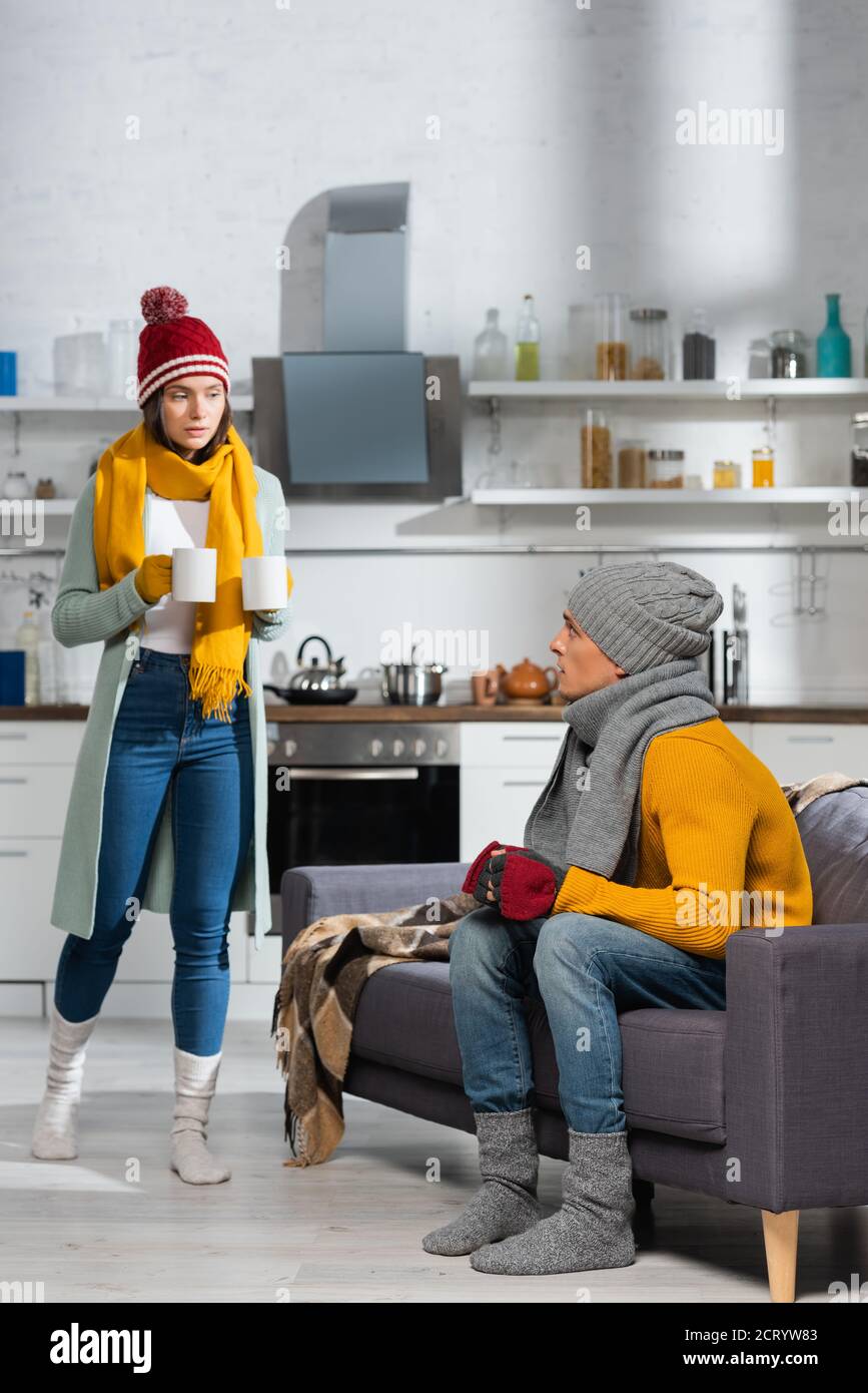 Frau in warmer Kleidung mit Tassen wärmende Getränke während Freund sitzt auf dem Sofa in der kalten Küche Stockfoto