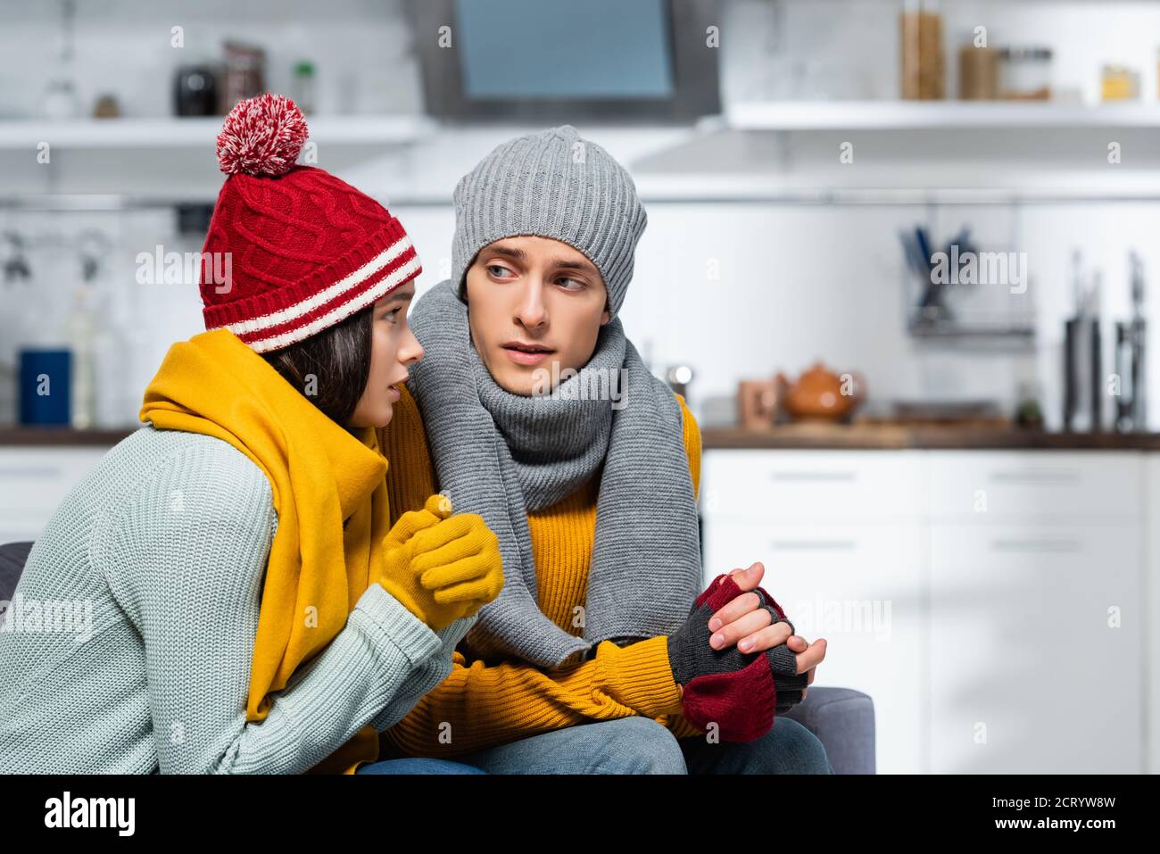 Junges Paar in gestrickten Hüten, Schals und Handschuhen, die sich beim Einfrieren in der kalten Küche angucken Stockfoto