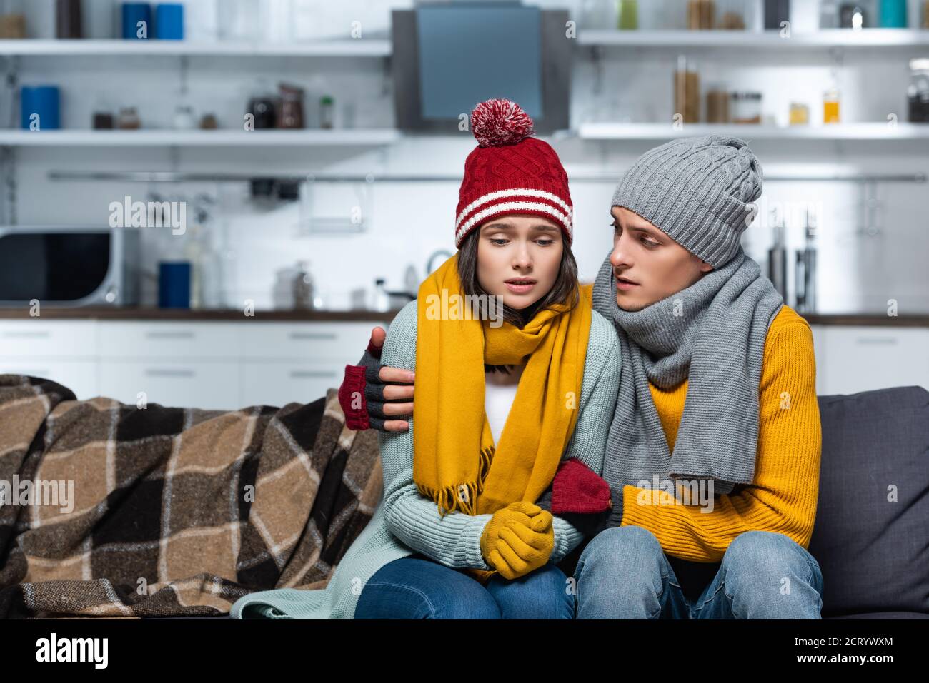 Junger Mann in warmen Hut und Schal umarmt eiskalt Freundin Während Sie in der kalten Küche sitzen Stockfoto