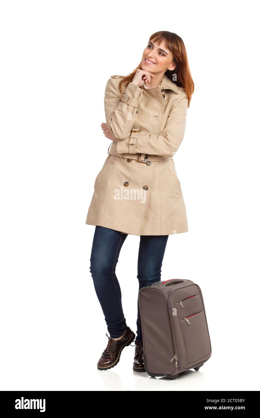Glückliche junge Reisende Frau in beigem Mantel, Jeans und braunen Schuhen steht neben einer Trolley-Tasche, hält Hand am Kinn, schaut weg und denkt. F Stockfoto