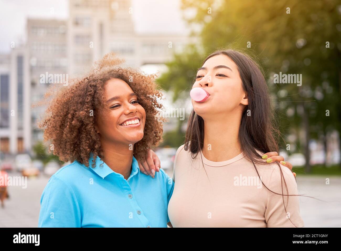 afroamerikanische Frau lachend, während asiatische Freund aufblasende Kaugummi. Nahaufnahme von multiethnischen Freunden, die die Straße im Freien genießen. Mädchen lachend an Stockfoto