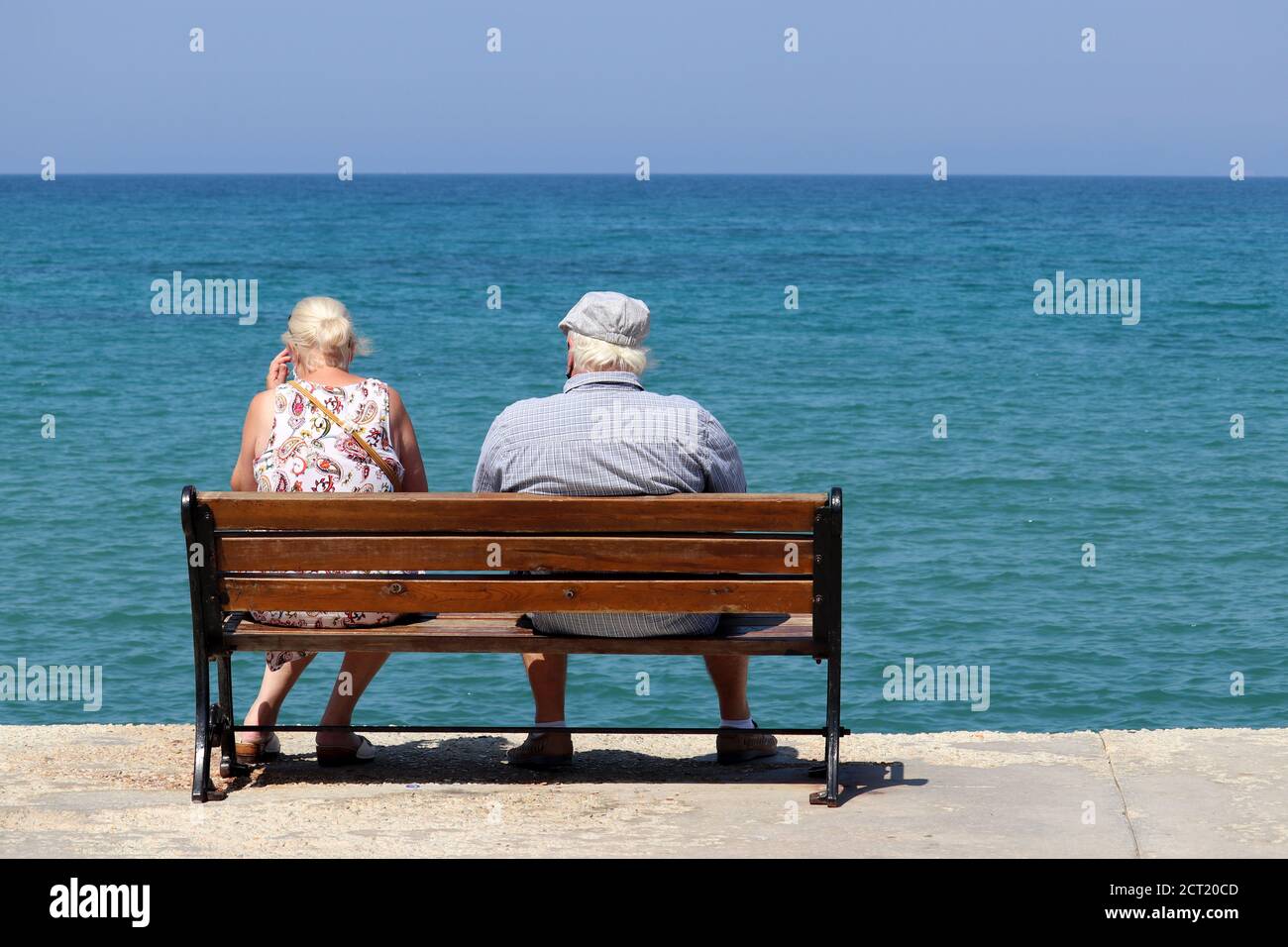 Ältere Frau und Mann sitzen auf einer Holzbank auf blauem Meeresgrund, Rückansicht. Altes Paar am Strand, entspannen und genießen das Leben im Ruhestand Stockfoto