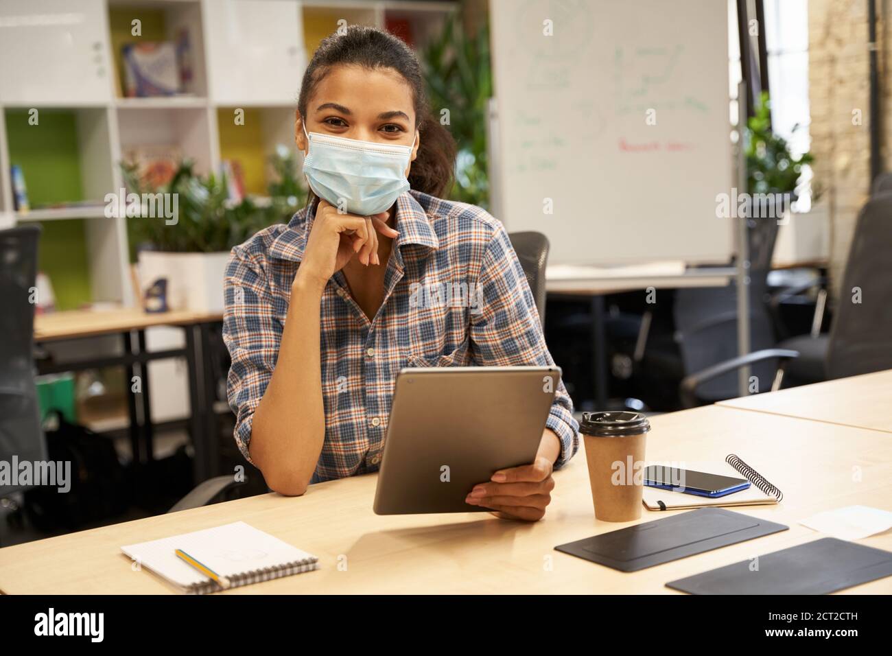 Junge Frau, weibliche Büroangestellte trägt eine schützende Gesichtsmaske, hält ein digitales Tablet und schaut auf die Kamera, während sie im modernen Büro arbeitet. Business und covid 19 Ausbruch, soziale Distanzierung Stockfoto