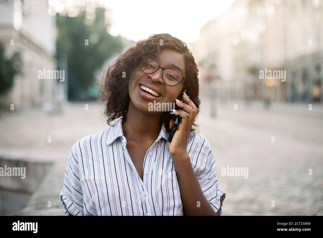 Nahaufnahme Porträt von attraktiven lockigen afrikanischen Mädchen reden auf dem Handy, Blick auf Kamera und lachen, zu Fuß im Freien auf der alten Stadtstraße an Stockfoto
