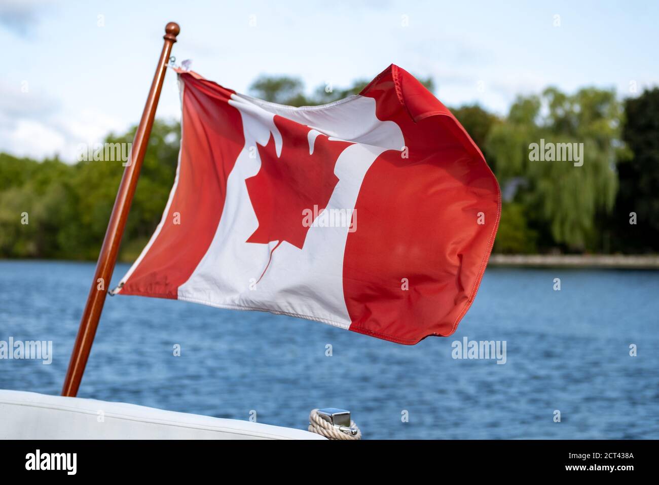 Eine kanadische Flagge fliegt im Wind am Heck oder Rücken eines kleinen Bootes. Bäume auf einem freien. Ufer und das blaue Wasser eines Flusses sind hinter ihm zu sehen. Stockfoto