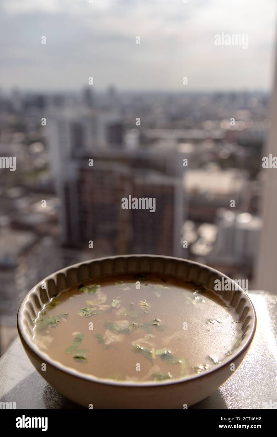 Hühnerbrühe mit Koriander in weißer Schale über Marmortisch mit Blick auf die Stadt am Morgen. Selektiver Fokus. Stockfoto