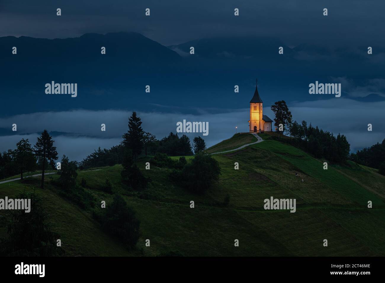 Jamnik, Slowenien - Blaue Stunde in Jamnik mit beleuchteter St. Primoz-Kirche auf einem Hügel in einem nebligen Sommeranbruch. Die Julischen Alpen im Hintergrund Stockfoto