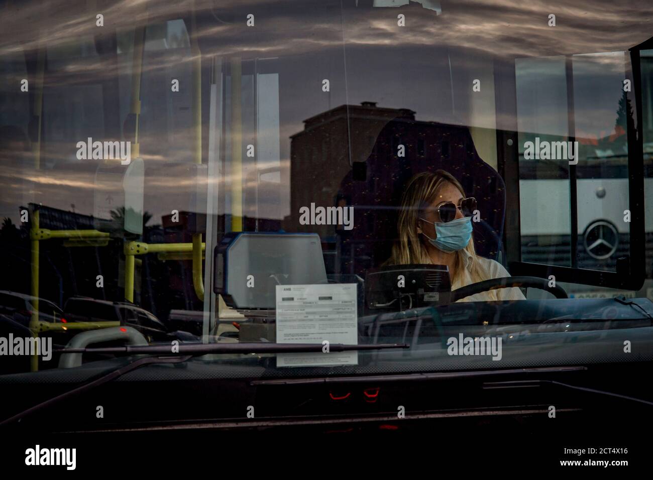 Der Himmel spiegelt sich auf dem Glas eines Stadtbusses in Barcelona, während der Fahrer, der eine Schutzmaske trägt, darauf wartet, dass die Ampel grün wird. Stockfoto