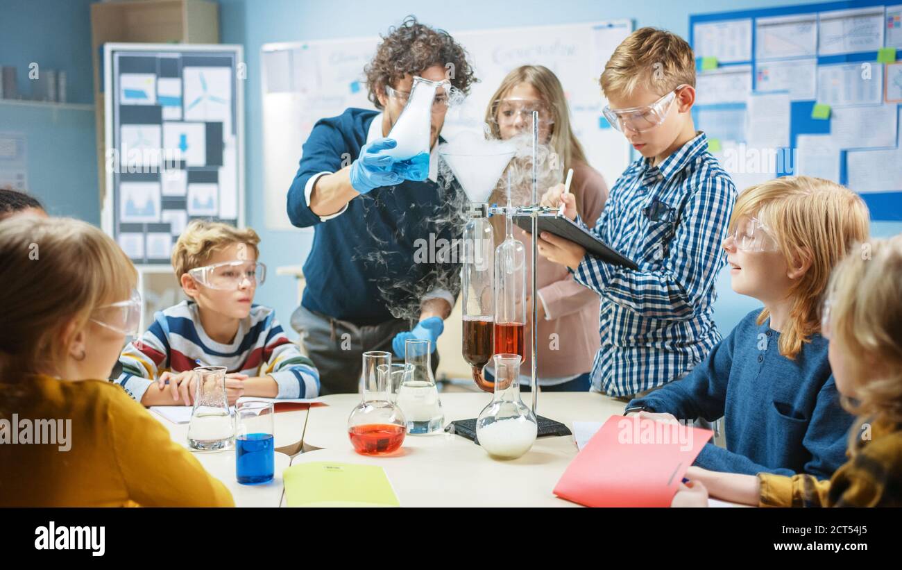 Grundschule Wissenschaft Klassenzimmer: Begeisterter Lehrer erklärt Chemie zu verschiedenen Gruppe von Kindern, zeigt ihnen, wie man Chemikalien in Bechern mischen Stockfoto