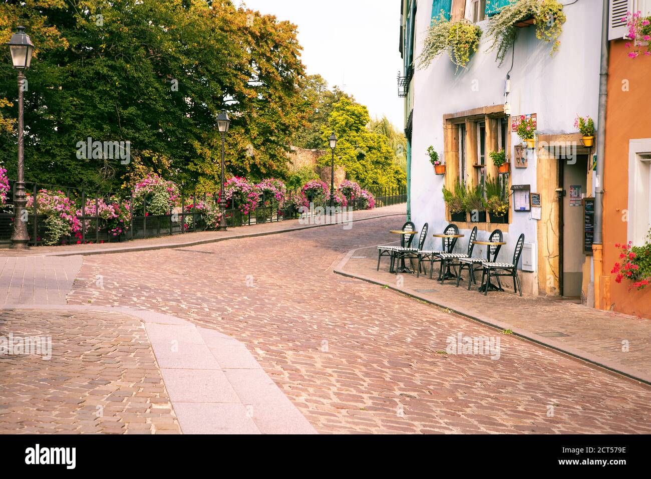 Blick auf romantische französische Kopfsteinpflasterstraße mit Café, Straßenlampen und Park im Hintergrund. Leere Tische und Stühle vor dem Restaurant Colmar Stockfoto