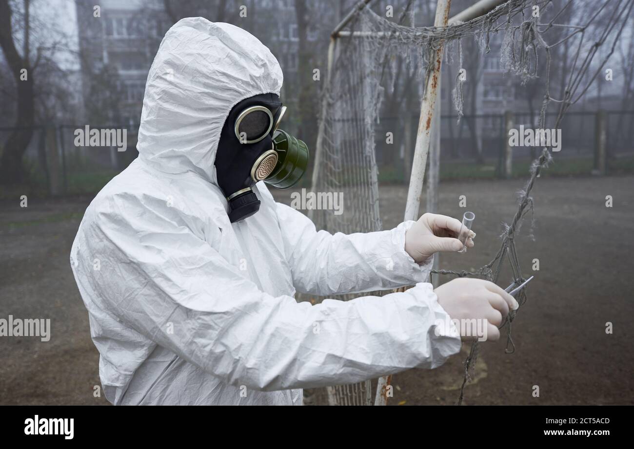 Seitenansicht des Ökologen im Strahlenanzug, der Pinzette und Reagenzglas hält, während die Proben auf verlassenen Straßen entnommen werden, Schutzkleidung, Gasmaske und Handschuhe tragen. Konzept der Umweltverschmutzung. Stockfoto