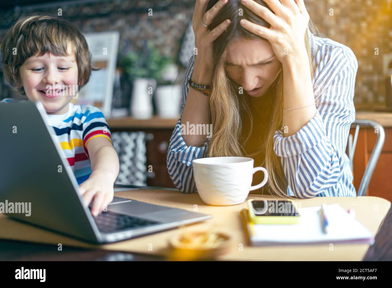 Mutter arbeitet von zu Hause und Online-Bildung mit Kind. Gestresst mit Computer und Telefon. Kind machen Lärm und stören Frau bei der Arbeit. Heimschooling und Stockfoto