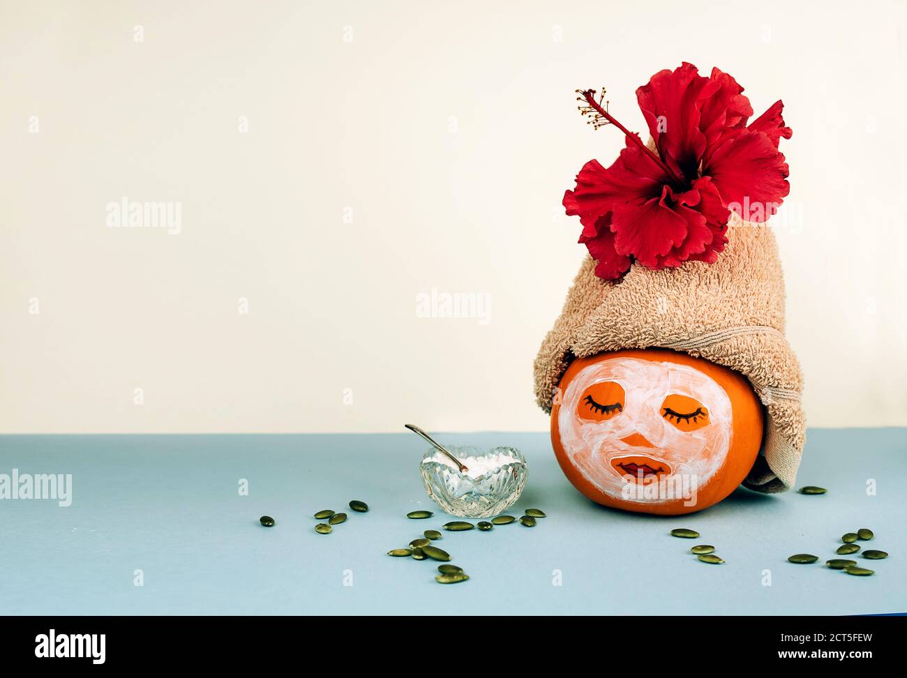 Kürbis mit bemaltem Gesicht und Gesichtsmaske, Handtuch mit Blume, Kürbiskernen und Puder in Glasschale auf pastellblauem Hintergrund. Spa-Konzept, Helloween, Kopierbereich. Stockfoto