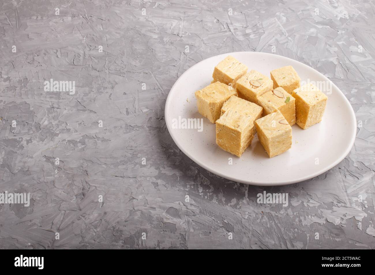 Traditionelle indische Süßigkeiten soan Papdi in weißem Teller mit Mandel und Pistasche auf einem grauen Betongrund. Seitenansicht, Kopierraum. Stockfoto