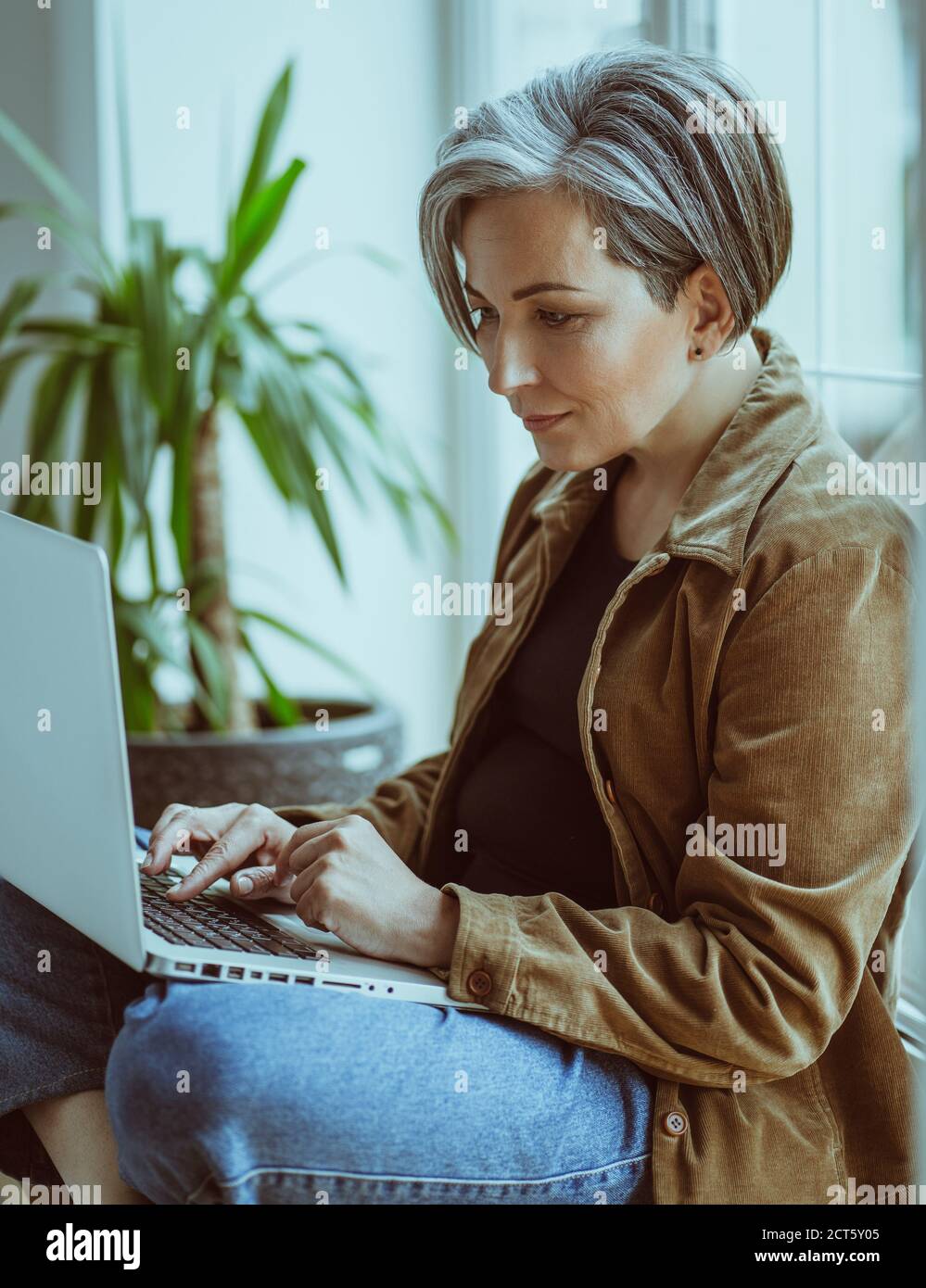 Elegante ältere Frau arbeitet Laptop auf Fensterbank sitzen. Seitenansicht der modernen silberhaarigen Lady in Casual Tippen auf Computer-Tastatur Stockfoto