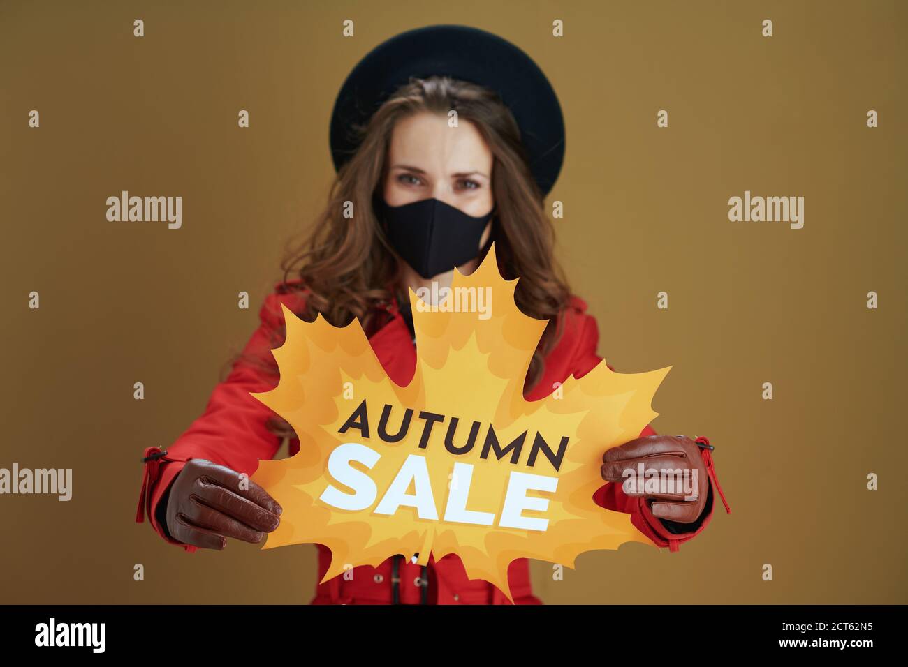 Hallo Herbst. Nahaufnahme einer Frau mittleren Alters im roten Mantel mit Herbstverkauf Blatt Form Banner und schwarze Maske auf braunem Hintergrund. Stockfoto