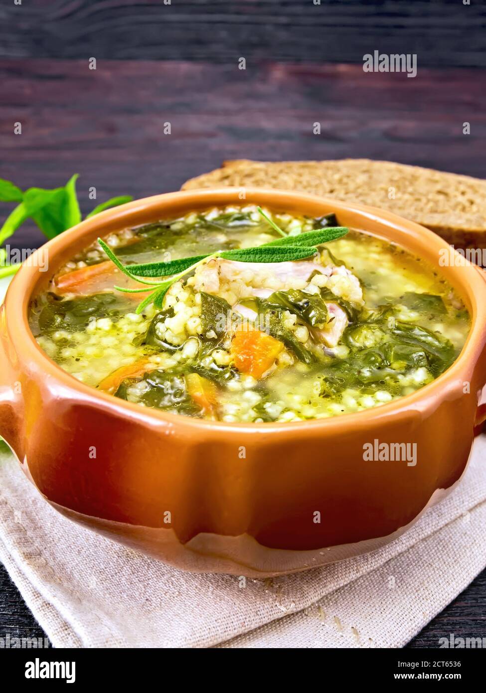 Schweinebrippen-Suppe mit Couscous und Spinat in einer Tonschüssel auf Handtuch, Brot gegen dunkles Holzbrett Stockfoto