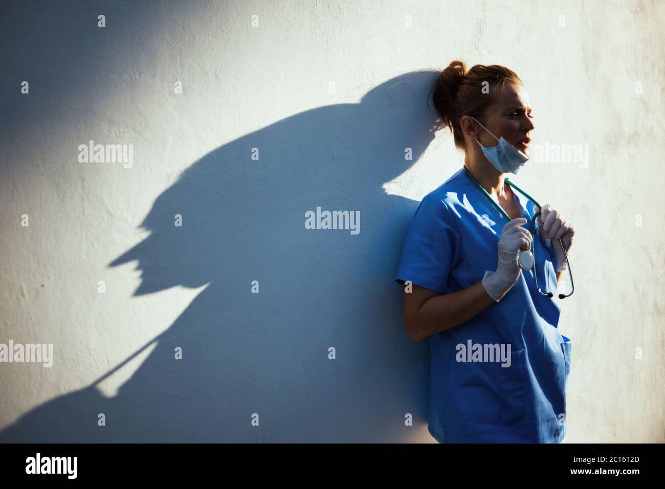 Covid-19 Pandemie. Müde moderne Arzt Frau in Scrubs mit Stethoskop, medizinische Maske und Gummihandschuhe außerhalb der Wand. Stockfoto