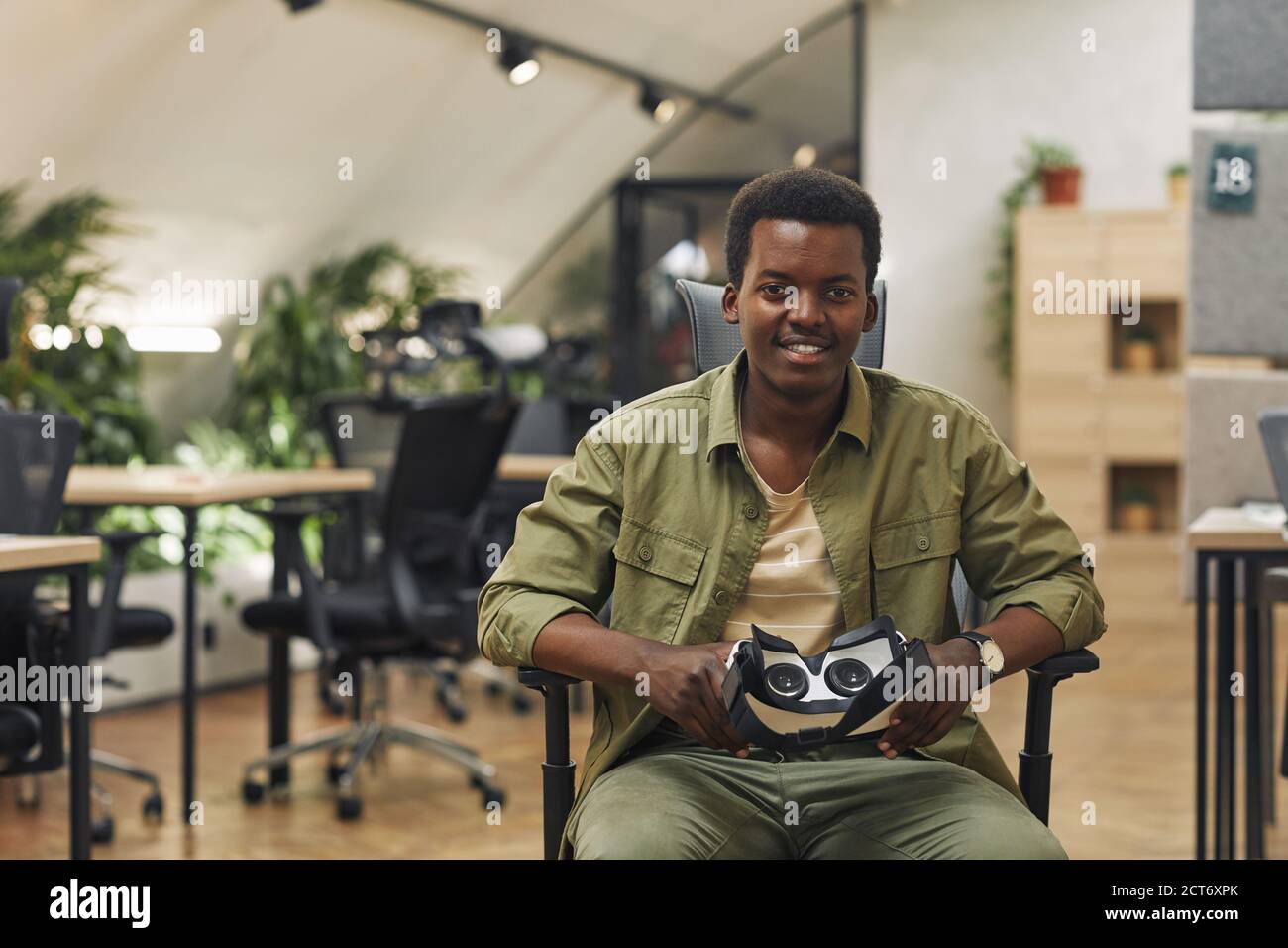 Porträt eines jungen afroamerikanischen Mannes, der VR-Ausrüstung hält und die Kamera im modernen Büro anlächelt, Kopierraum Stockfoto