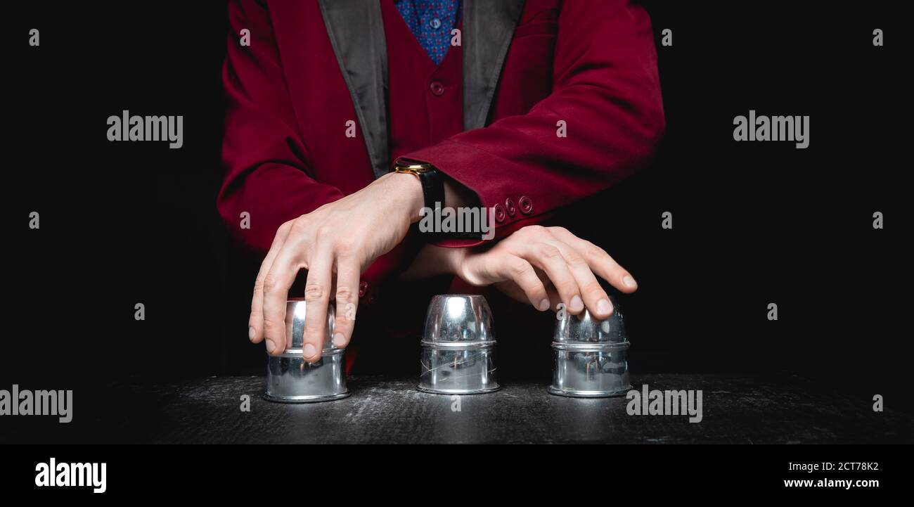 Magier zeigt Schalenspiel von Thimbeln mit Kreisen und Ball, schwarzer Hintergrund. Konzept Täuschung, Schlittenhand Stockfoto
