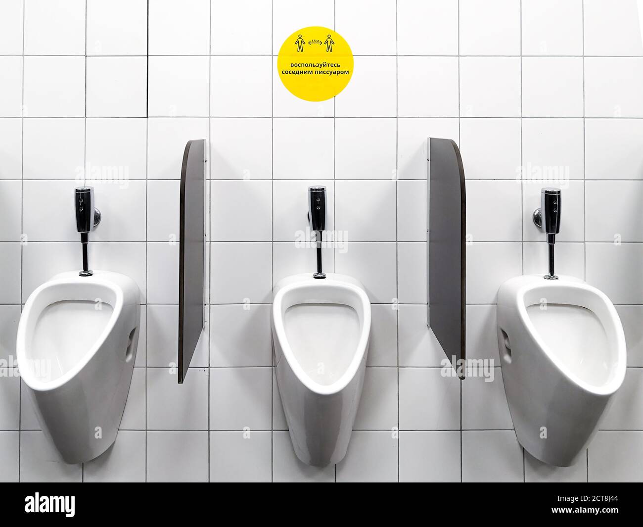 Urinale an der weißen Wand einer öffentlichen Männertoilette. Ein gelbes Zeichen mit Text auf Russisch fördert soziale Distanz. Vorbeugende Maßnahmen zu schützen Stockfoto