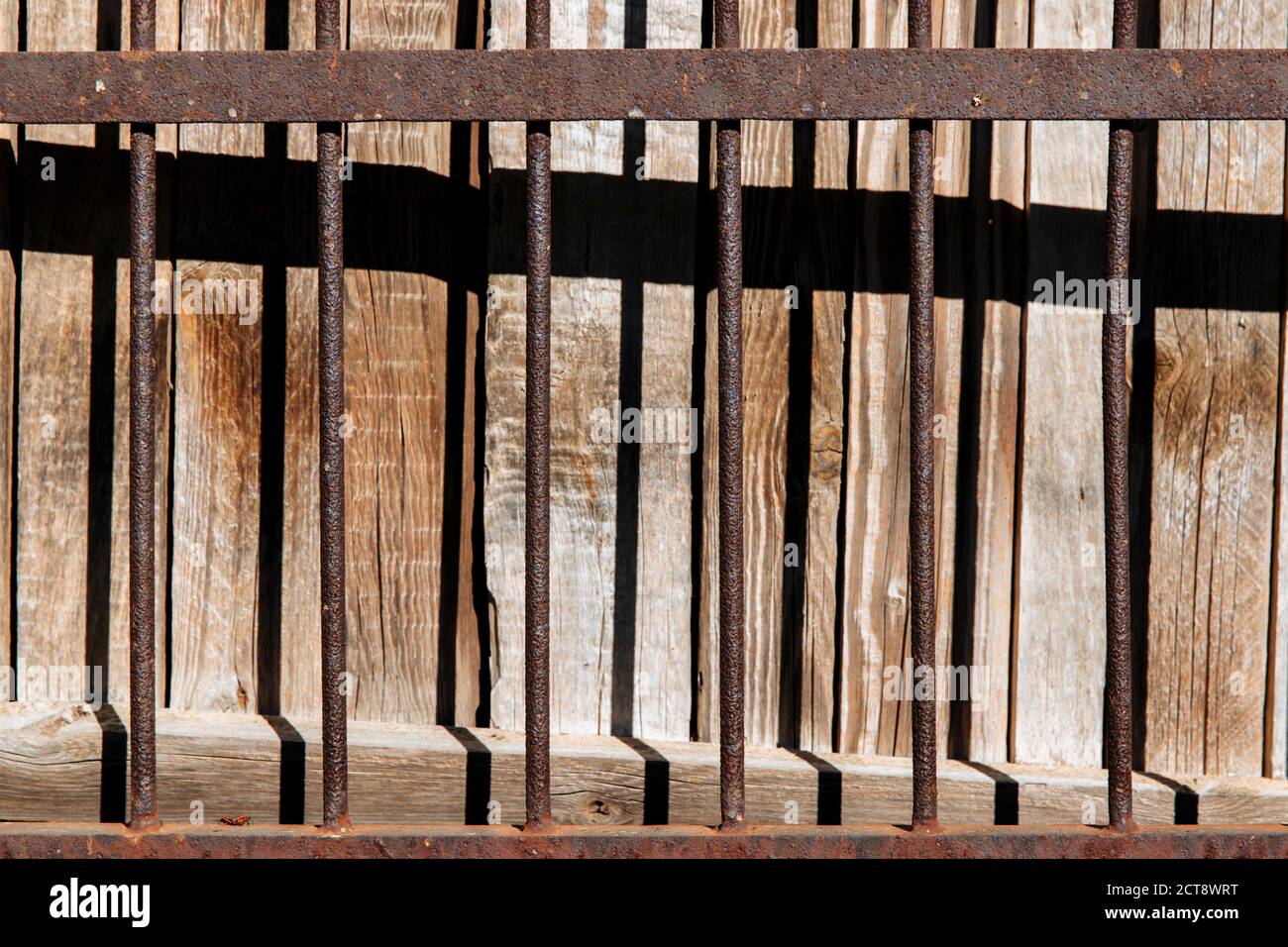 Ein alter, rostig geschmiedeter Zaun. Die Textur eines massiven Metallzauns. Hinter dem Zaun befindet sich ein Holzhintergrund, auf den der Schatten fällt. Stockfoto