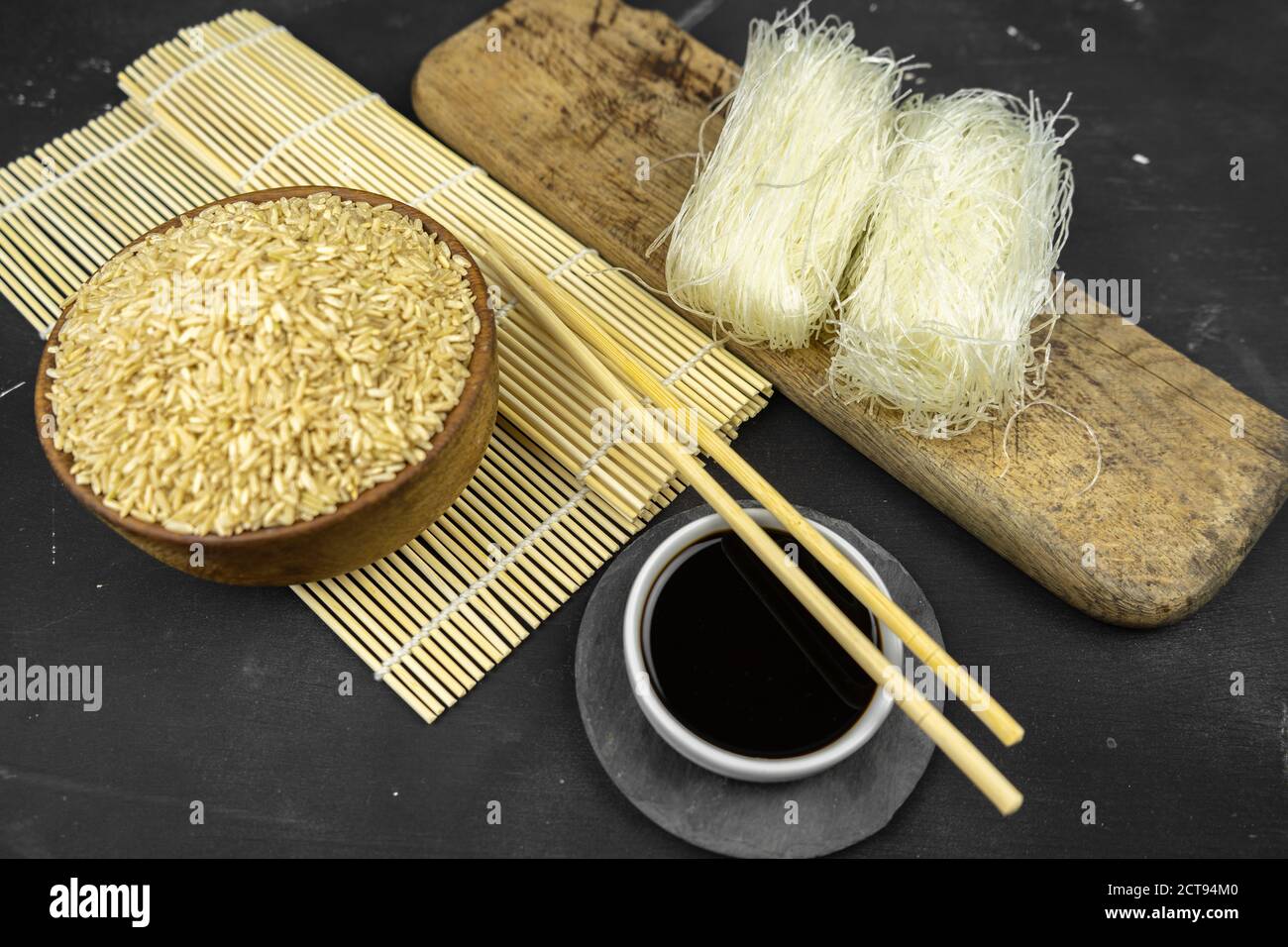 Brauner Reis in einer Holzschüssel, asiatischer Küchenhintergrund mit Bambusmatte, Essstäbchen, Sojasauce, eingelegtem Ingwer und Glasreis-Nudeln auf schwarzem Backgroun Stockfoto