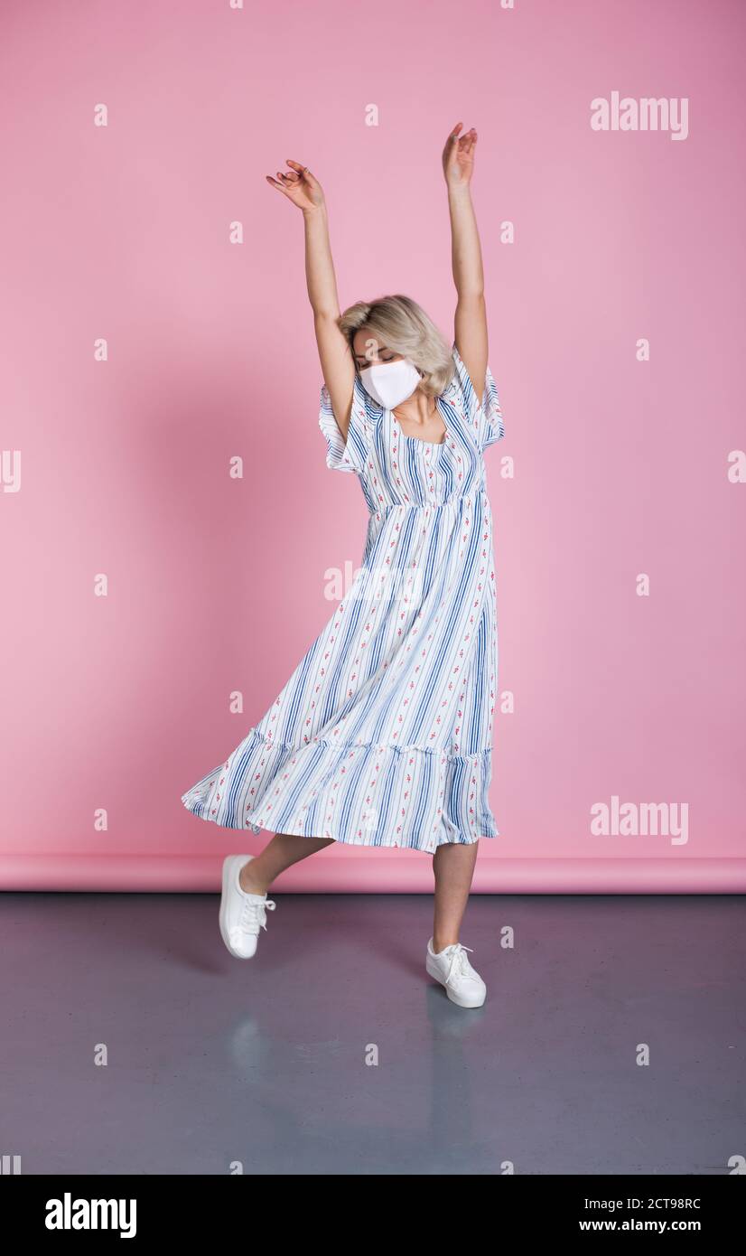 Charmante Frau im Sommerkleid trägt eine medizinische Maske auf Face tanzt auf einer rosafarbenen Studiowand Stockfoto