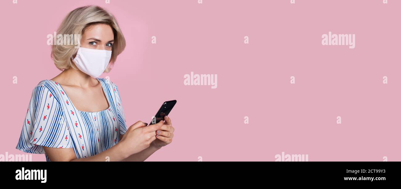 Blonde Frau Werbung etwas, während posiert auf einem rosa Studio Wand mit Freiraum und Maske auf Gesicht und tragen Halten eines Telefons Stockfoto