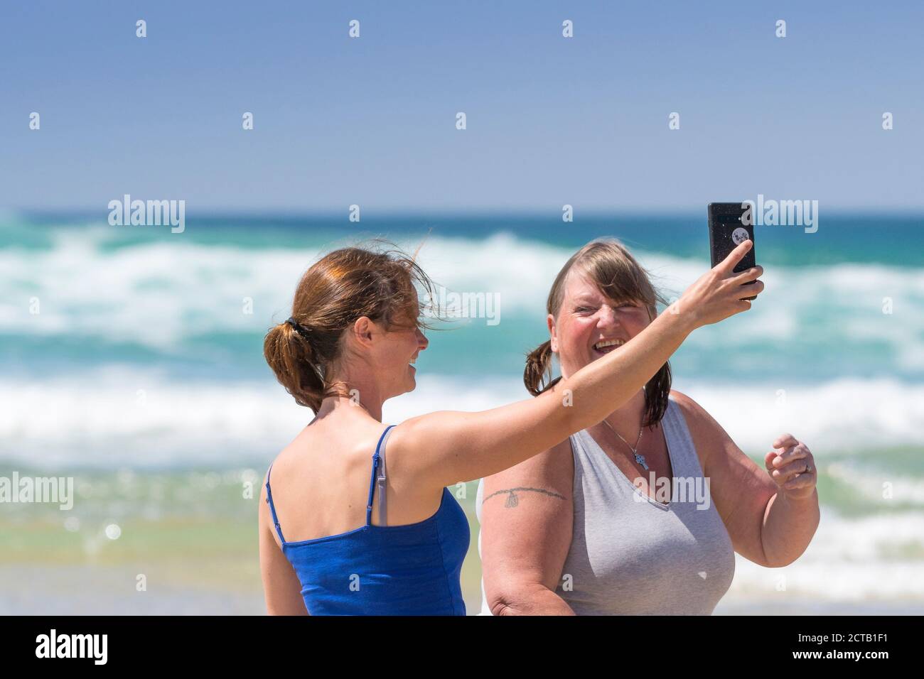 Frauen, die während ihres Aufenthaltes in den Urlaub fahren, lachen, als sie ein Selfie mit einem Smartphone am Fistral Beach in Newquay in Cornwall machen. Stockfoto