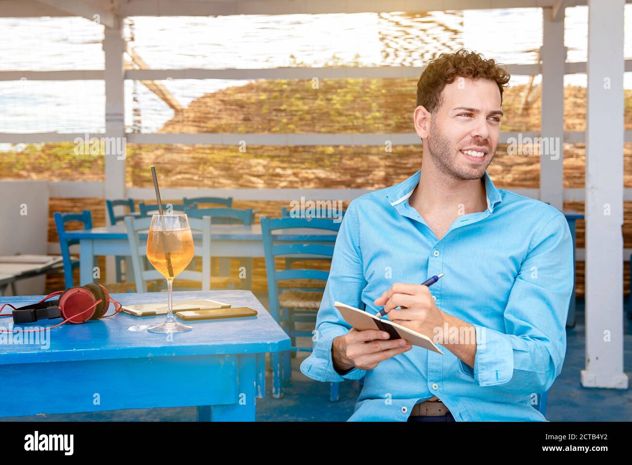 Linkshändiger junger Mann, der an der Strandbar sitzt und schreibt, was er tut Hausaufgaben Konzept der Smart-Working Outdoor Stockfoto