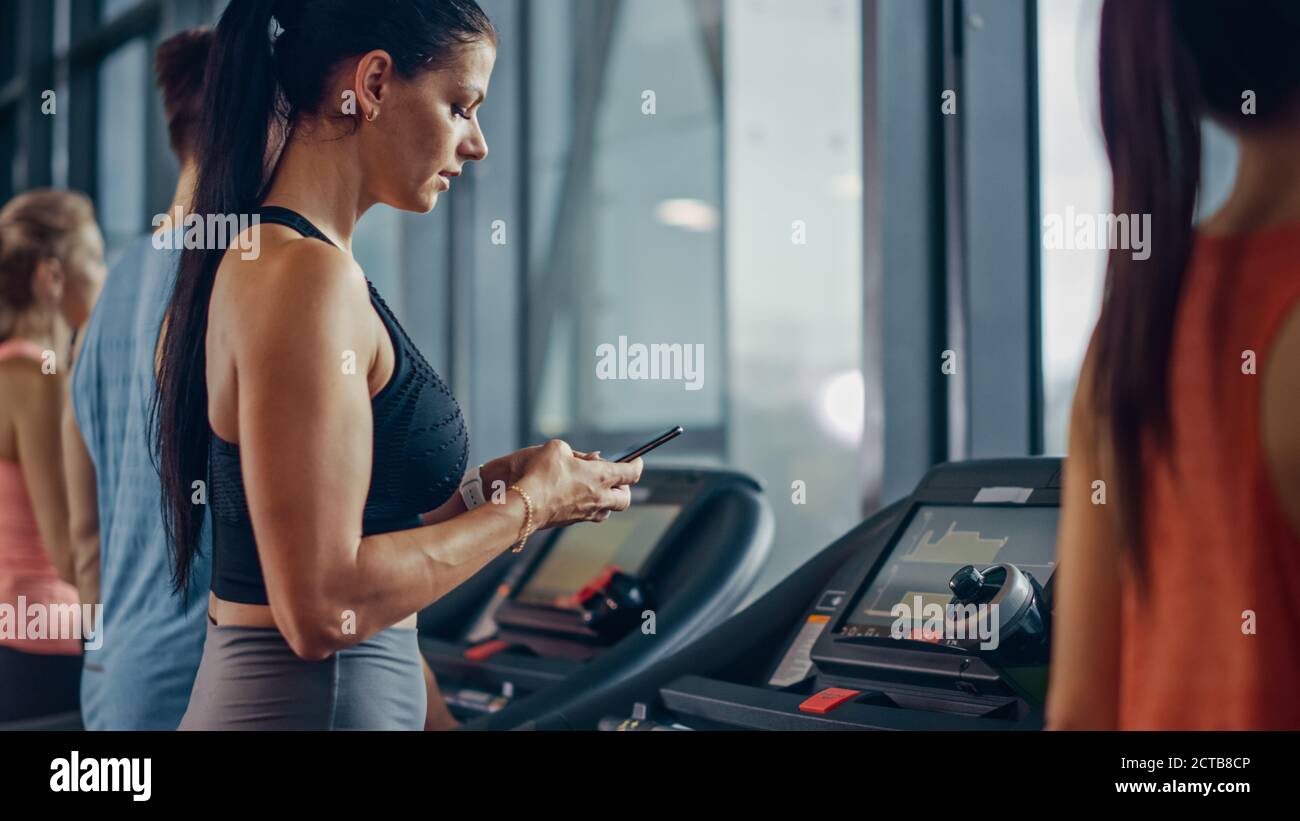 Schöne Athletic Sport Frau in der Turnhalle läuft auf einem Laufband, nutzt Smartphone. Im Hintergrund Fit Athleten Training. Seitenansicht Stockfoto