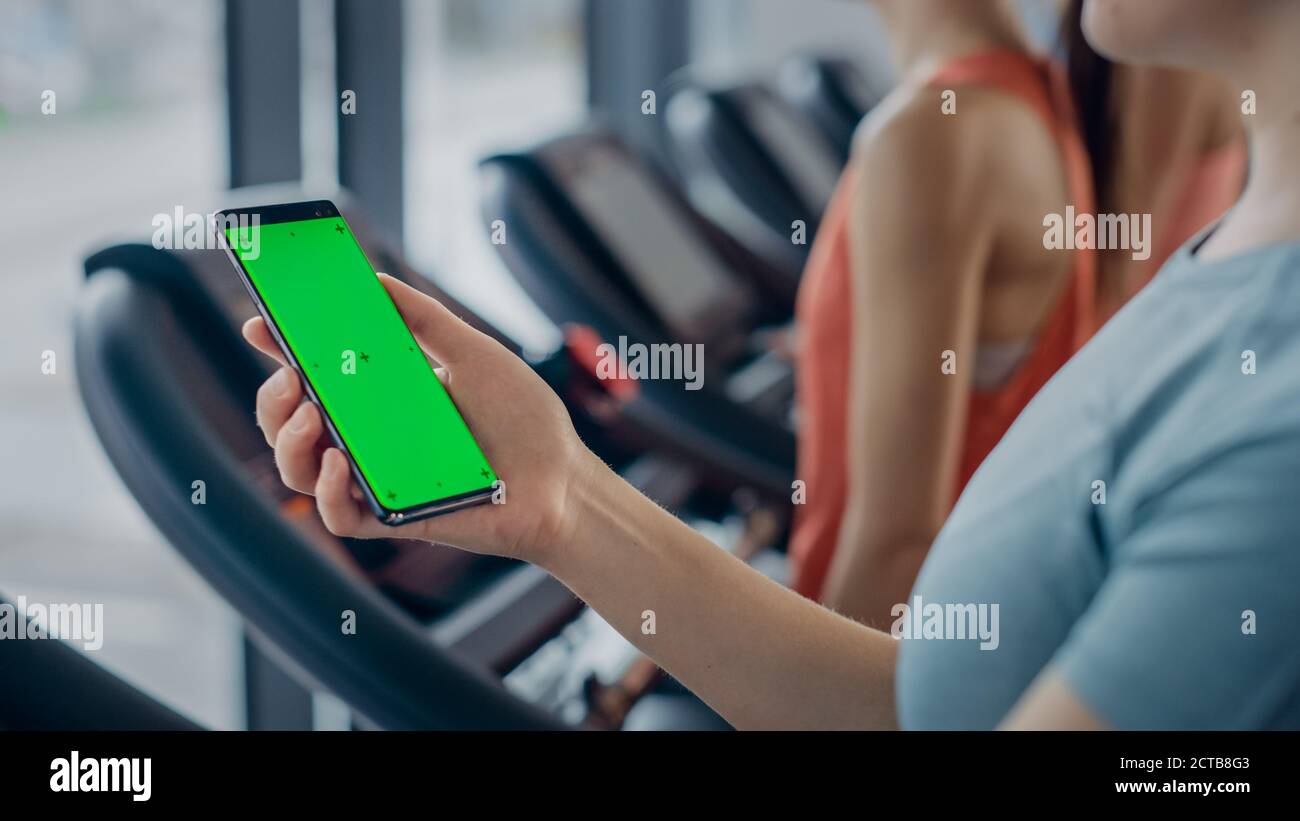 Athletische Frau verwendet Green Mock-up Bildschirm Smartphone beim Laufen auf dem Laufband in einem Fitnessstudio. Sport weibliche Beiträge auf Social Media, nimmt Bilder Stockfoto